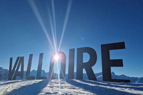 Schneebedeckter Boden mit dem Schriftzug "VALLOIRE" vor einem klaren blauen Himmel und schneebedeckten Bergen im Hintergrund.