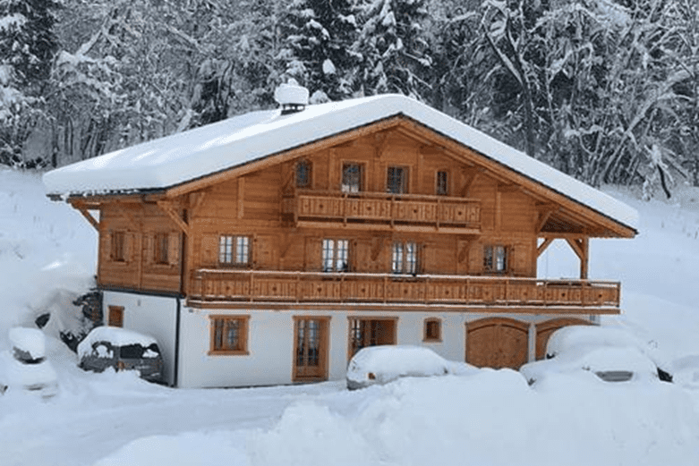 Holzhaus im Schnee mit zwei Etagen und Balkonen; umgeben von verschneiten Bäumen und parkenden Autos.