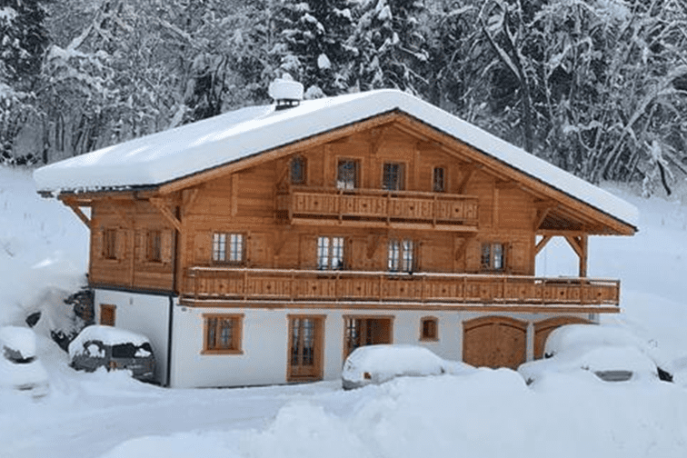 Holzhaus im Schnee mit zwei Etagen und Balkonen; umgeben von verschneiten Bäumen und parkenden Autos.