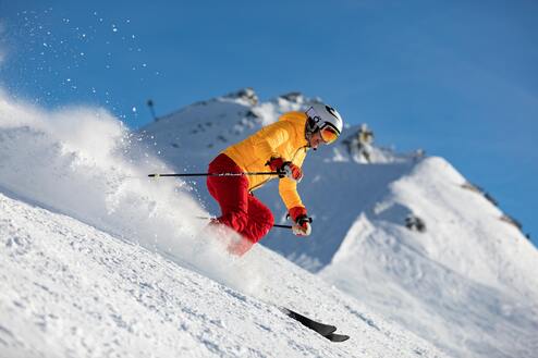 Skifahrer in gelber Jacke und roten Hosen fährt eine Piste hinunter, Schnee spritzt in alle Richtungen.
