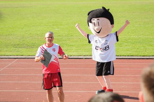 Mann in Sportkleidung spricht auf einem Sportplatz neben einem gro&szlig;en Maskottchen in Fu&szlig;balltrikot.