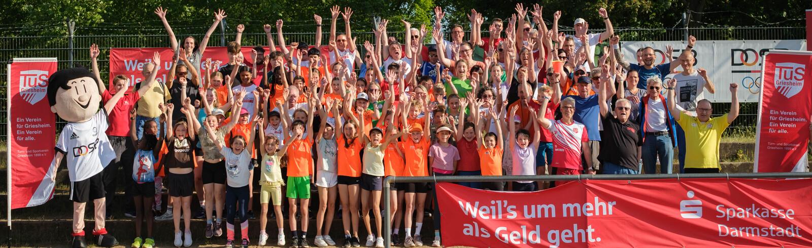 Gruppenfoto von Teilnehmern auf einem Sportgel&auml;nde, l&auml;chelnd mit erhobenen H&auml;nden, umgeben von Sponsorenbanner.