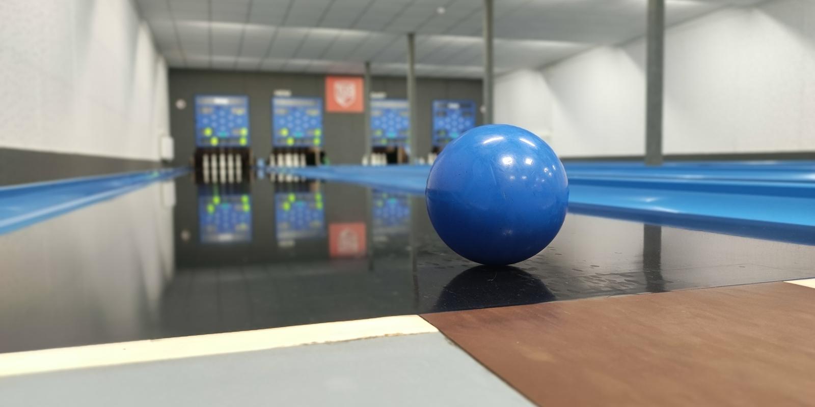 Blauer Bowlingball liegt auf der Bahn, mit Anzeigetafeln und Pins im Hintergrund.