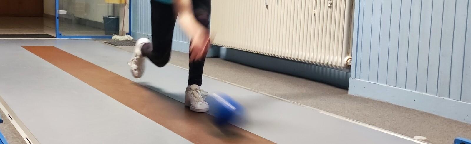 Eine Person in Sportkleidung rollt eine blaue Kugel auf einer Bowlingbahn. Im Hintergrund sind Fenster und Heizk&ouml;rper sichtbar.