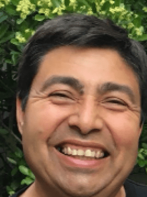 Lächelnder Mann vor grünem Hintergrund mit Pflanzen, trägt ein schwarzes T-Shirt.