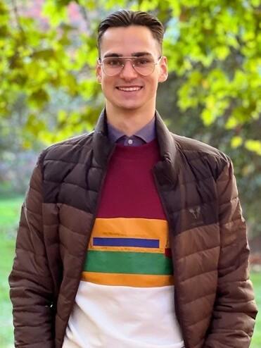 Junger Mann mit Brille in warmer Jacke und buntem Pullover steht lächelnd in einem parkähnlichen Umfeld mit Bäumen.