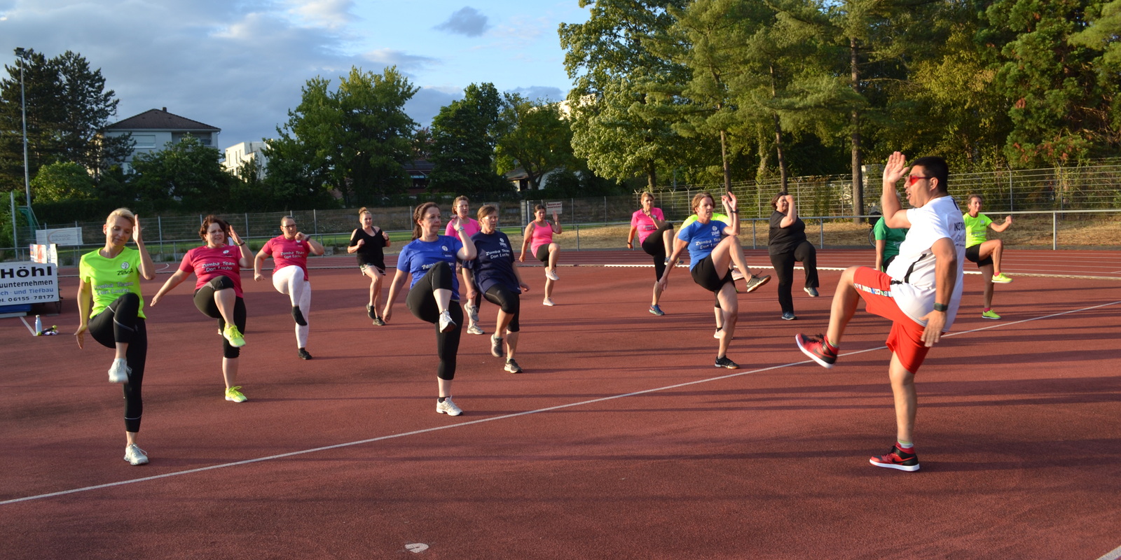 Gruppe von Frauen und einem Mann in Sportbekleidung beim Aufw&auml;rmen auf einem roten Sportplatz unter bew&ouml;lktem Himmel.