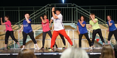 Zwei Männer und sechs Frauen in Sportkleidung tanzen synchron auf einer Bühne mit einem Geländer im Hintergrund.