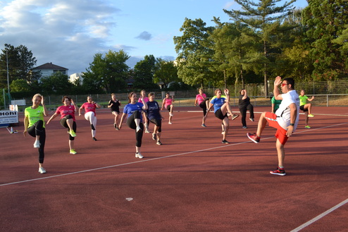 Gruppe von Frauen und einem Mann in Sportbekleidung beim Aufw&auml;rmen auf einem roten Sportplatz unter bew&ouml;lktem Himmel.