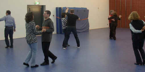 Tanzende Paare in einem hellen Raum mit blauen Bodenbel&auml;gen, verschiedene Tanzstile erkennbar.