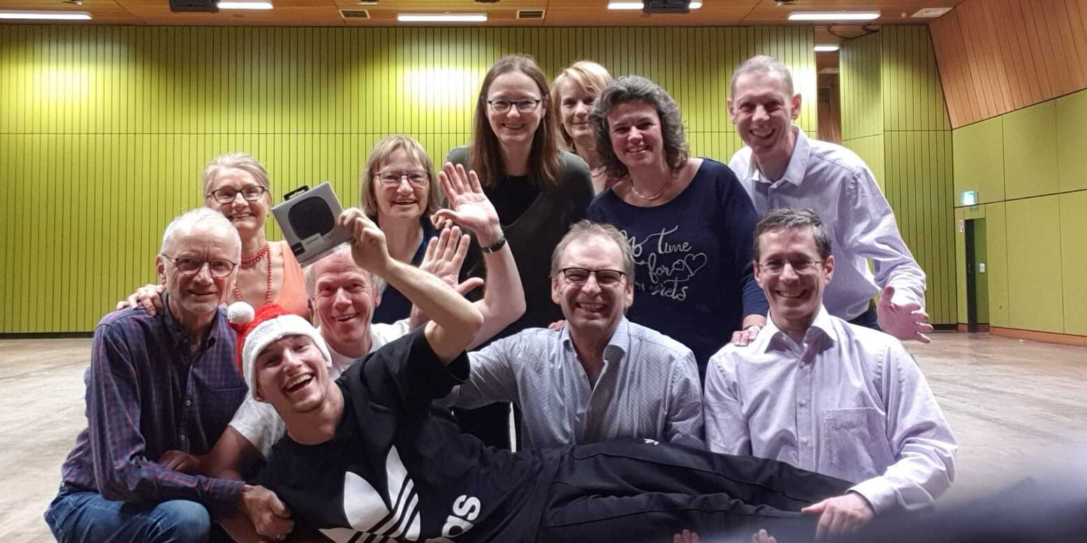 Gruppenfoto von zw&ouml;lf Personen, die l&auml;cheln und verschiedene Gesten im Innenraum eines Veranstaltungssaals zeigen.