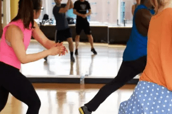 Gruppe von T&auml;nzern in einem Spiegelsaal, die synchron zu Musik eine Choreografie vorf&uuml;hren.