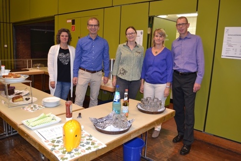Fünf Personen stehen an einem Tisch mit Snacks und Getränken in einem Raum mit grünen Wänden.