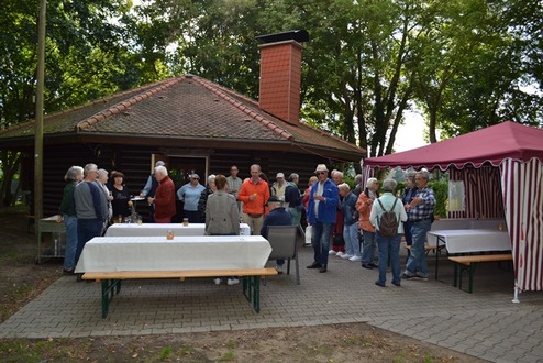 Gruppierung von Personen vor einer Holzlaube, umgeben von Bäumen, mit Sitzgelegenheiten und einem Pavillon im Hintergrund.