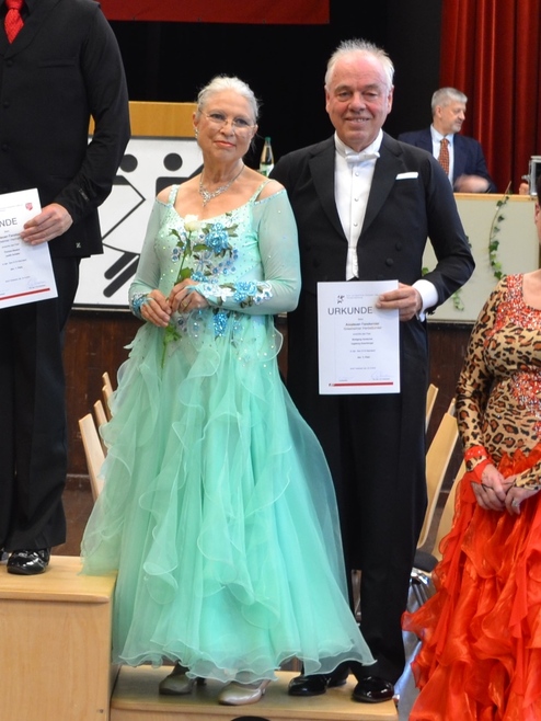 Tanzpaar in eleganter Kleidung hält Urkunden und Blumen auf einer Veranstaltung. Rote und grüne Kleider sind sichtbar.