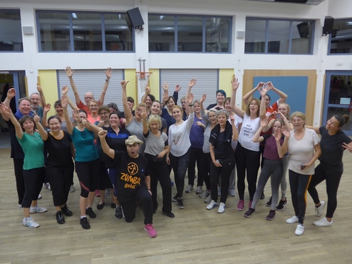 Gruppenszene mit vielen Menschen in Sportkleidung, die begeistert zu einer Zumba-Stunde posieren und feiern.