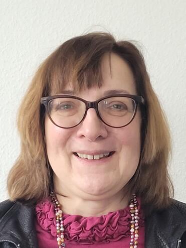 Portr&auml;t einer l&auml;chelnden Frau mit Brille, blonder Frisur, schwarzer Lederjacke und buntem Halskette vor hellem Hintergrund.