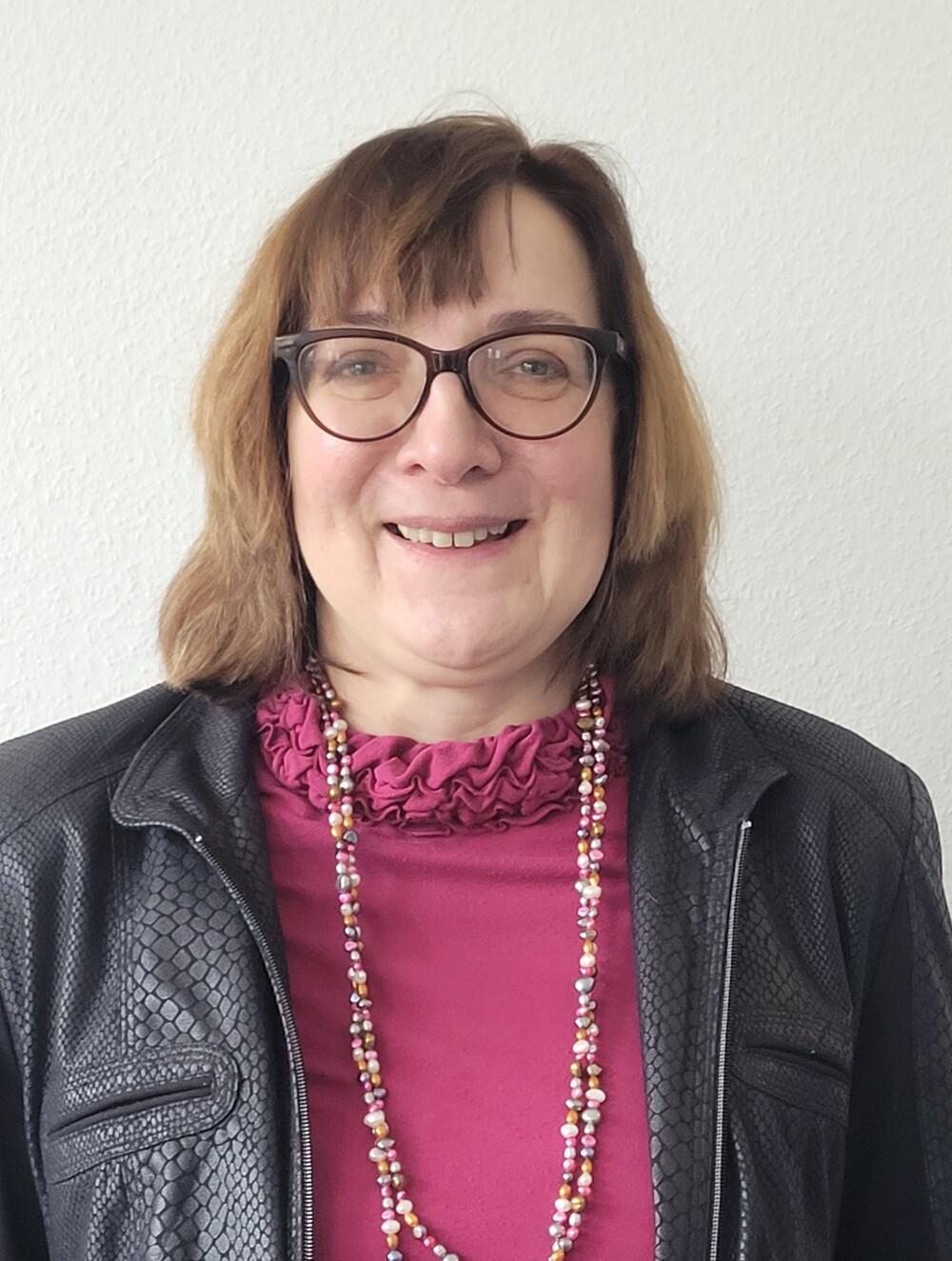 Portr&auml;t einer l&auml;chelnden Frau mit Brille, blonder Frisur, schwarzer Lederjacke und buntem Halskette vor hellem Hintergrund.