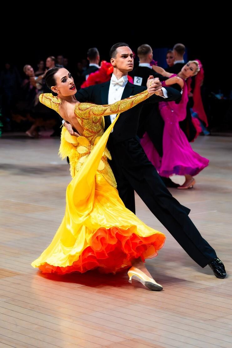 Tanzpaar in eleganter Bewegung; die T&auml;nzerin tr&auml;gt ein gelbes Kleid mit orangefarbenem Saum, der T&auml;nzer einen schwarzen Anzug.