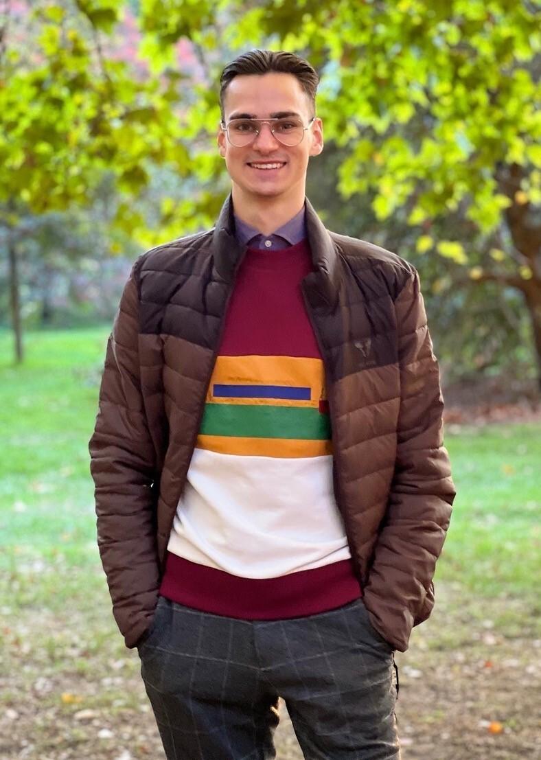 Junger Mann mit Brille in warmer Jacke und buntem Pullover steht lächelnd in einem parkähnlichen Umfeld mit Bäumen.