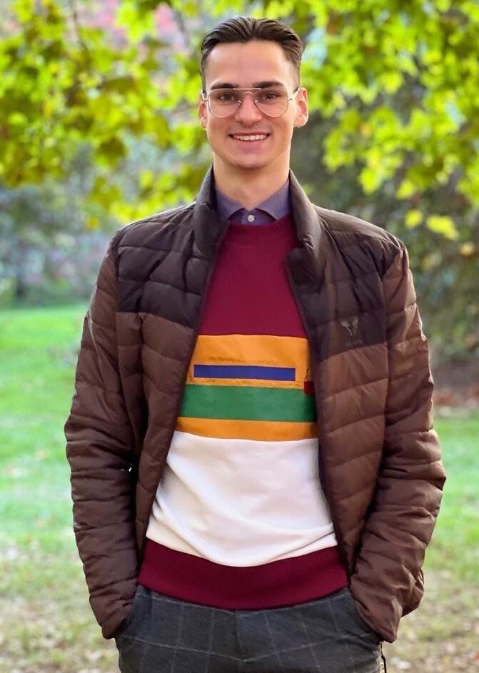 Junger Mann mit Brille in warmer Jacke und buntem Pullover steht l&auml;chelnd in einem park&auml;hnlichen Umfeld mit B&auml;umen.