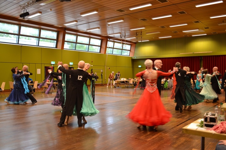 Tanzpaaren in eleganter Abendgarderobe tanzen in einer hellen Sporthalle mit grünen Wänden und großen Fenstern.