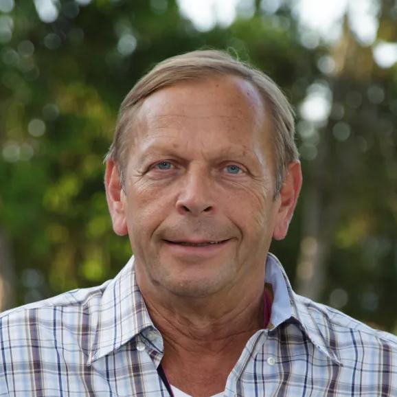 Lächelnder Mann mit blauen Augen in kariertem Hemd vor einem verschwommenen, grünen Hintergrund.