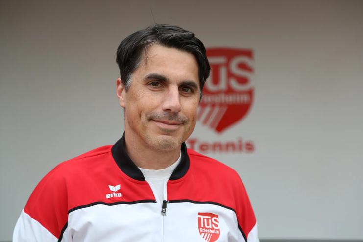 Portr&auml;t eines Mannes in Sportjacke mit Vereinslogo vor einem Hintergrund mit dem Schriftzug &bdquo;TUS Gr&uuml;nstein Tennis&ldquo;.