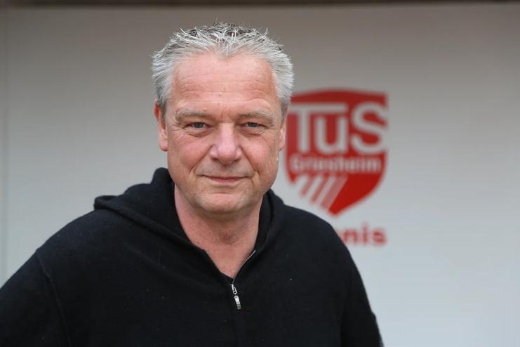 Portrait eines Mannes mit grauen Haaren, der in einem schwarzen Hoodie vor einer Wand mit dem Logo von TuS Gr&auml;fenhain steht.