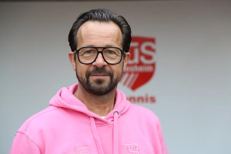 Mann mit Brille und Bart tr&auml;gt ein pinkes Sweatshirt vor einer Wand mit einem Tennis-Logo.
