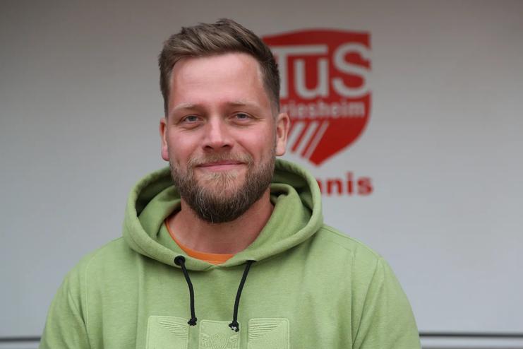 Portr&auml;t eines l&auml;chelnden Mannes mit Bart, der einen gr&uuml;nen Hoodie tr&auml;gt, vor einem Hintergrund mit dem Logo des TuS.