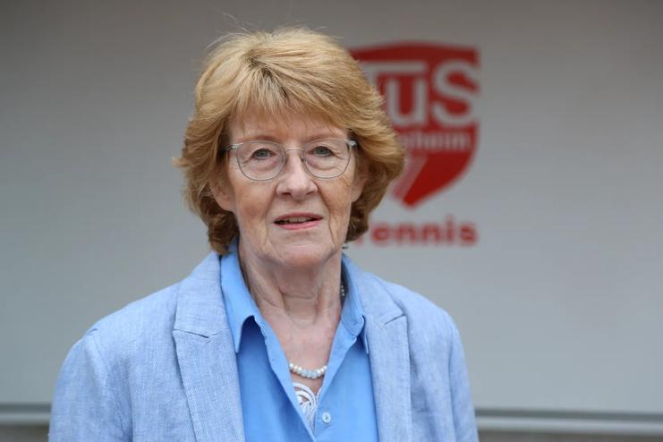 Portr&auml;t einer Frau mit blonden, lockigen Haaren, die in einem blauen Blazer vor einem Tischtennis-Schild steht.