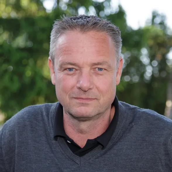 Nahaufnahme eines Mannes mit kurzen Haaren, in einem grauen Pullover, vor einem unscharfen Hintergrund aus Bäumen.