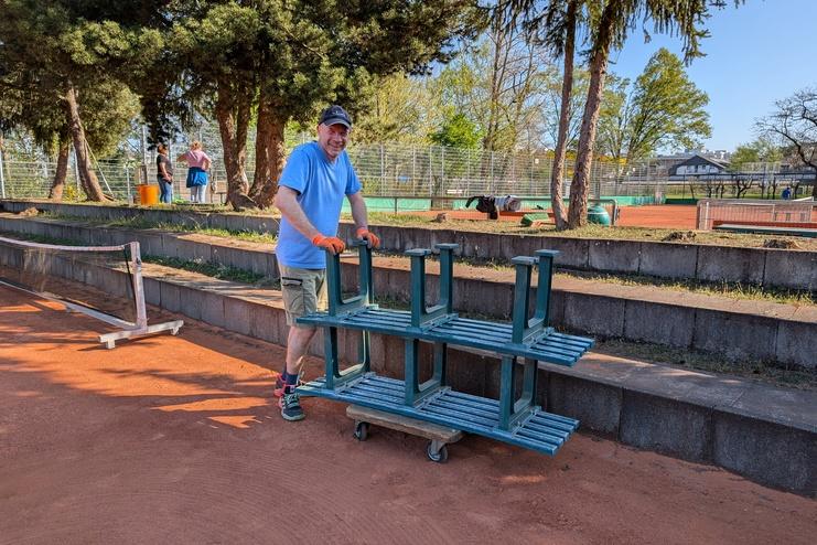 Mann schiebt ein Gestell auf Rollen mit gestapelten B&auml;nken auf einem Tennisplatz mit gr&uuml;nen B&auml;umen im Hintergrund.