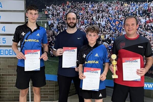 Vier Tischtennisspieler präsentieren Urkunden und einen Pokal vor einem bunten Hintergrund mit vielen Zuschauern.