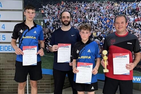 Vier Tischtennisspieler präsentieren Urkunden und einen Pokal vor einem bunten Hintergrund mit vielen Zuschauern.