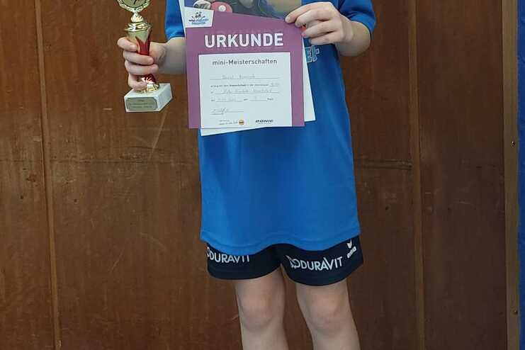 Junge in blauer Sportbekleidung hält Pokal und Urkunde in einer Sporthalle, steht auf einer Trainingsfläche.