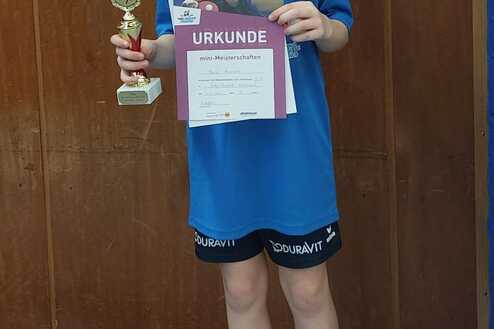 Junge in blauer Sportbekleidung hält Pokal und Urkunde in einer Sporthalle, steht auf einer Trainingsfläche.