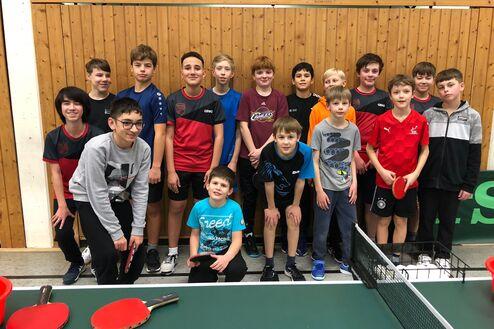 Gruppenshot von 16 Kindern in Sportkleidung vor einer Tischtennisplatte mit Schlägern und Bällen.