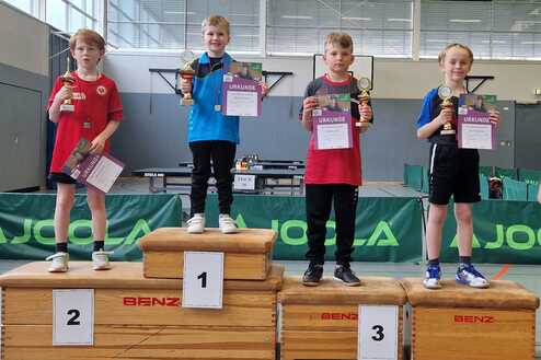 Vier Kinder stehen auf einem Siegertreppchen, halten Pokale und Urkunden in Händen, mit Tischtennisplatten im Hintergrund.