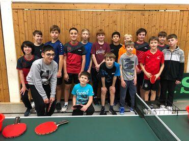 Gruppenshot von 16 Kindern in Sportkleidung vor einer Tischtennisplatte mit Schlägern und Bällen.