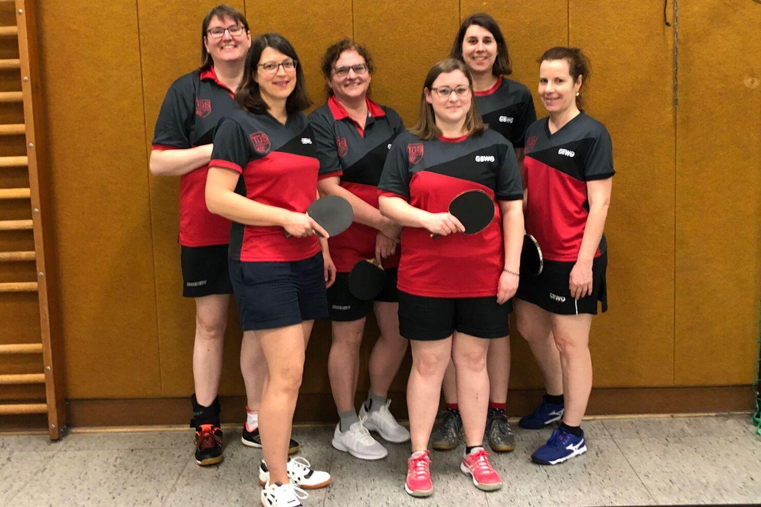 Sechs Frauen in sportlichen Outfits mit Tischtennisschlägern stehen lächelnd vor einer Wand in einer Sporthalle.