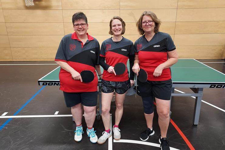 Drei Tischtennisspielerinnen in passenden Trikots stehen vor einem Tischtennistisch und halten Schläger in den Händen.