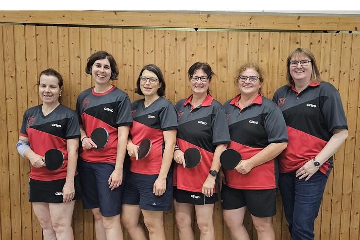 Sechs Frauen in Tischtenniskleidung posieren lächelnd mit Schlägern vor einer Holzwand.