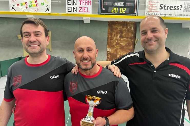 Drei Männer in Sporttrikots stehen zusammen und halten einen Pokal, im Hintergrund eine Tischtennisplatte und Banner.