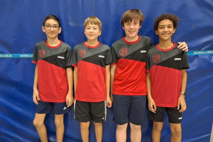 Vier Jungen in roten und grauen Sporttrikots stehen vor einem blauen Hintergrund.