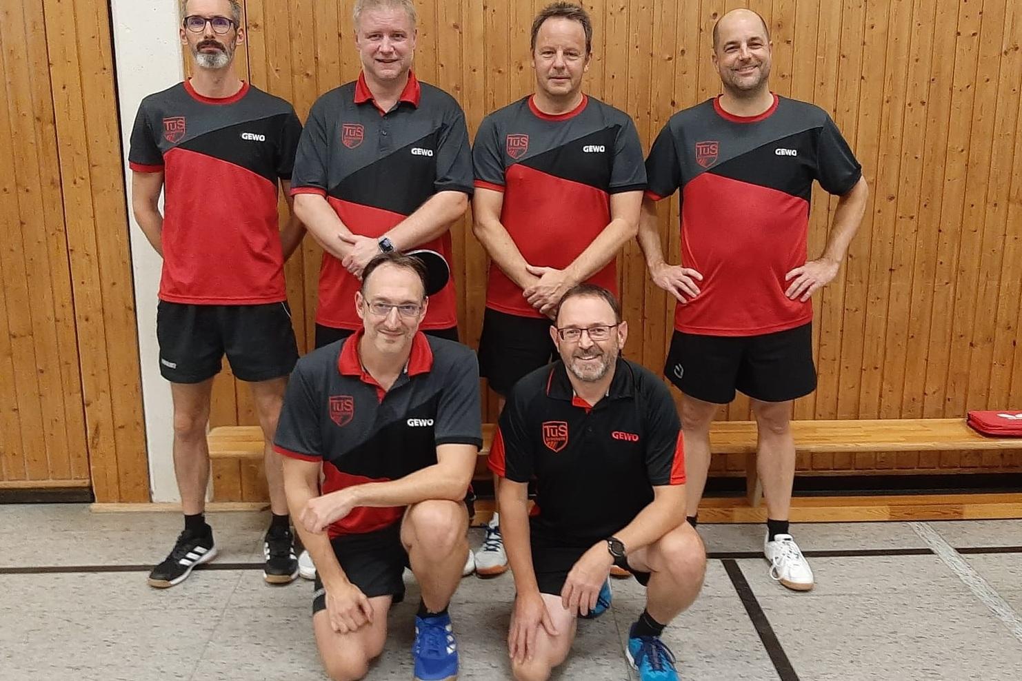 Sechs Tischtennisspieler in roten und schwarzen Trikots posieren in einer Sporthalle vor einer Holzwand.