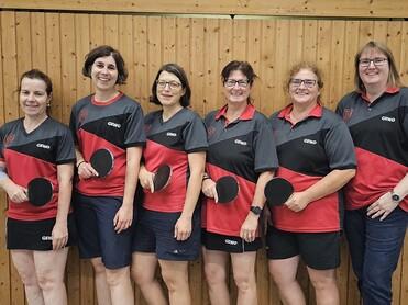 Sechs Frauen in Tischtenniskleidung posieren lächelnd mit Schlägern vor einer Holzwand.