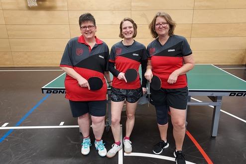 Drei Tischtennisspielerinnen in passenden Trikots stehen vor einem Tischtennistisch und halten Schläger in den Händen.