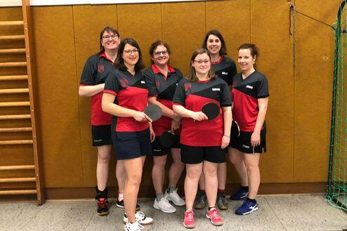 Sechs Frauen in sportlichen Outfits mit Tischtennisschlägern stehen lächelnd vor einer Wand in einer Sporthalle.